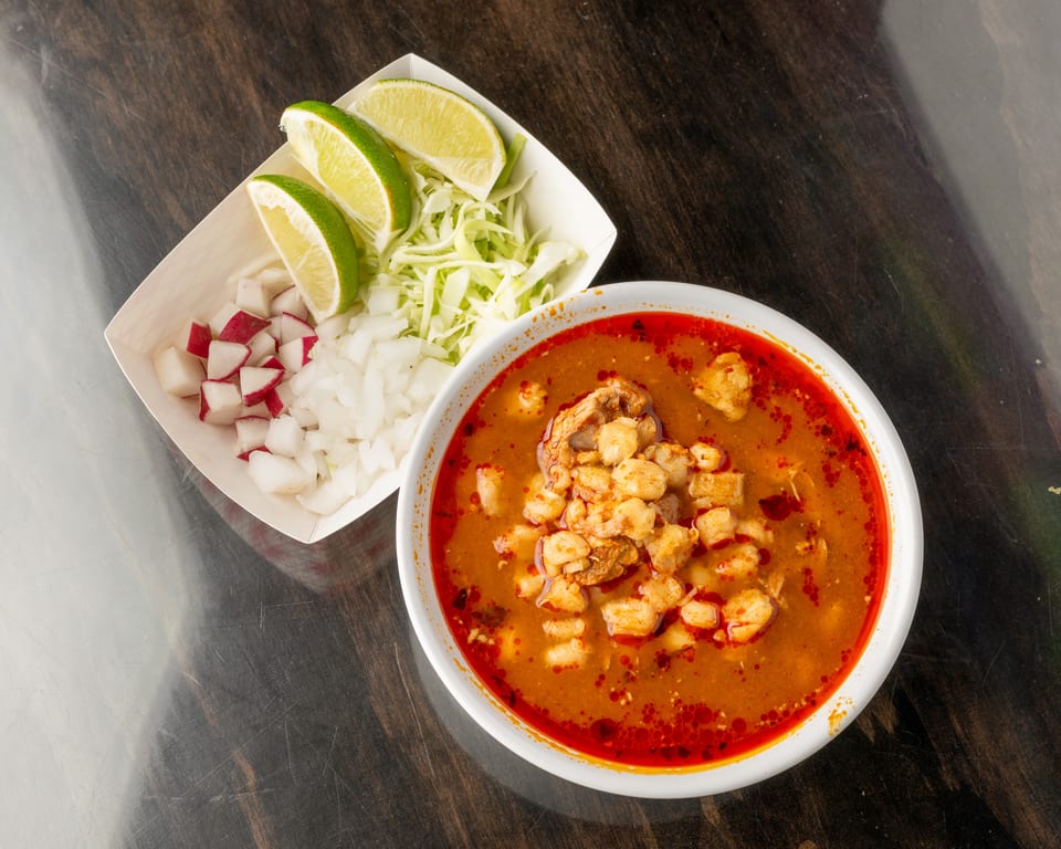 A bowl of pozole with a side of diced radishes, onions, shredded cabbage, and lime wedges.