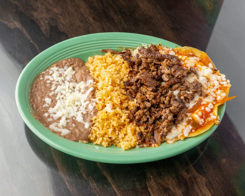 A green plate holds refried beans with cheese, yellow rice, and chopped meat over cheesy enchiladas.