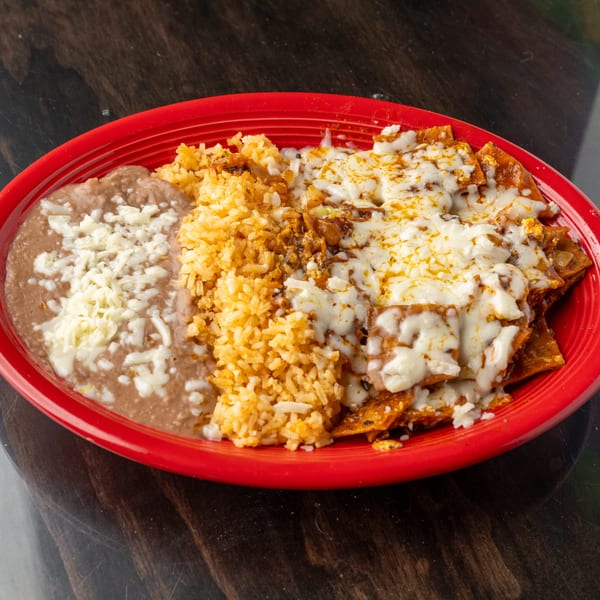 A red plate holds chilaquiles with melted cheese, refried beans, and Spanish rice on a dark table.