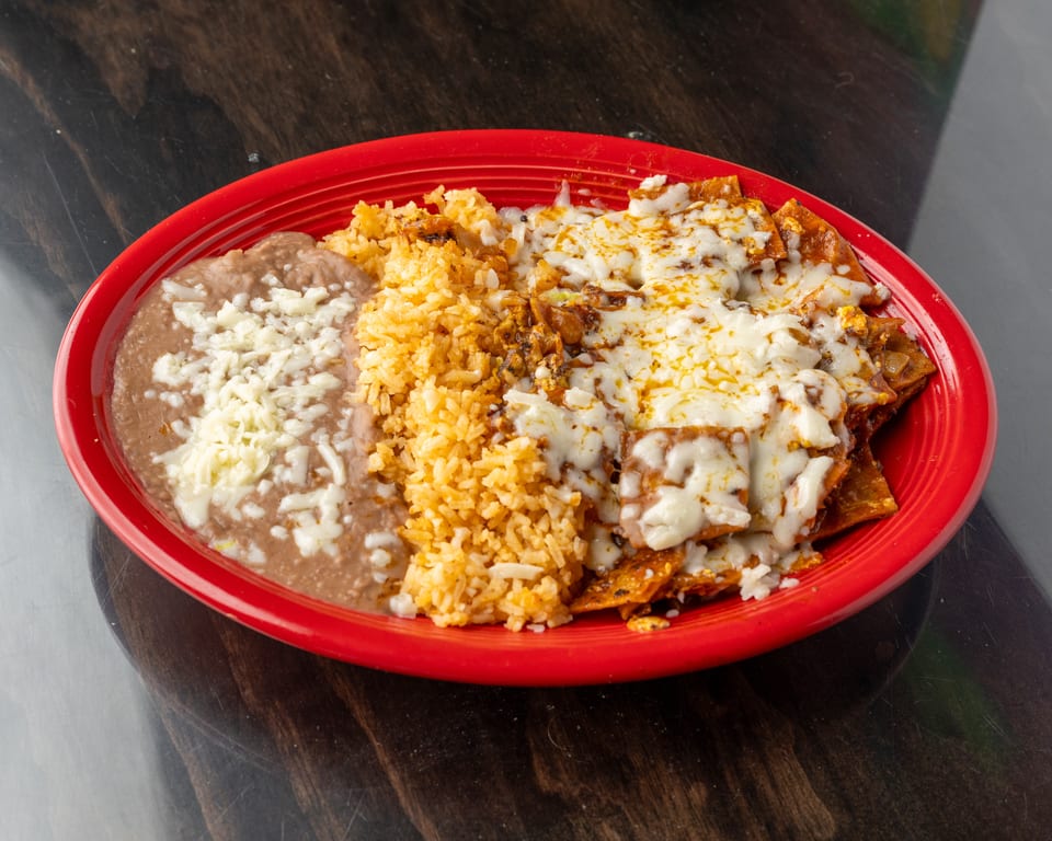 A red plate holds chilaquiles with melted cheese, refried beans, and Spanish rice on a dark table.