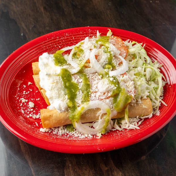 Crispy taquitos topped with green salsa, sour cream, shredded lettuce, onions, and cheese on a red plate.