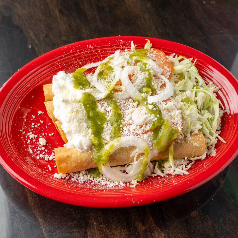 Crispy taquitos topped with green salsa, sour cream, shredded lettuce, onions, and cheese on a red plate.