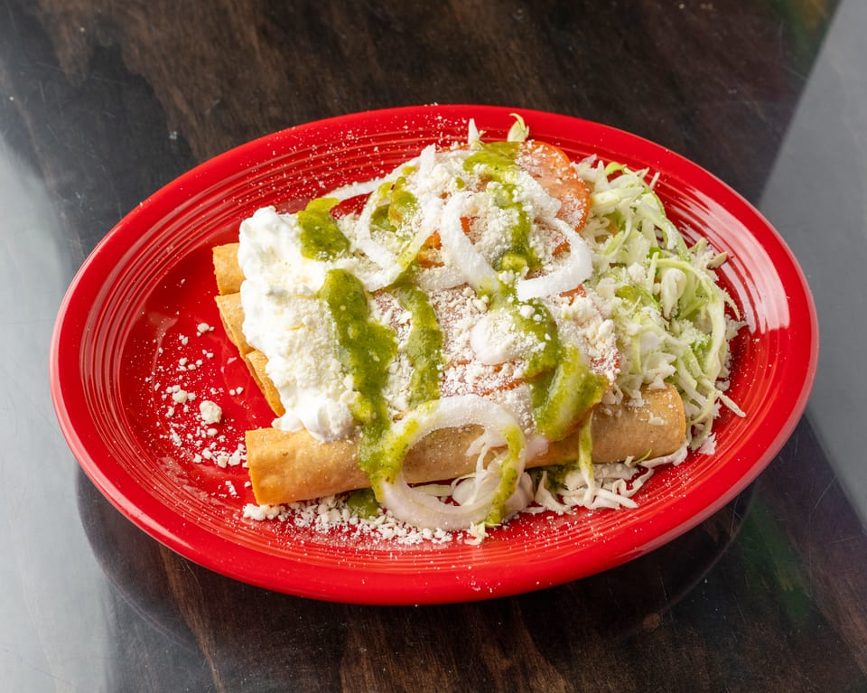 Crispy taquitos topped with green salsa, sour cream, shredded lettuce, onions, and cheese on a red plate.