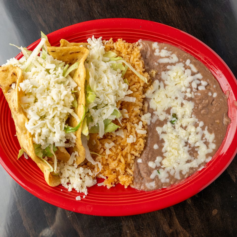 Two crispy tacos with lettuce and cheese, served with rice and refried beans on a red plate.