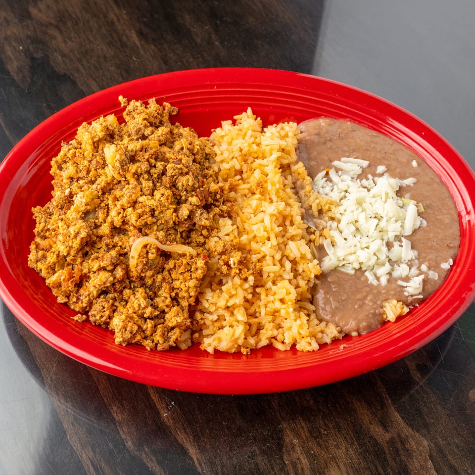 Scrambled eggs with chorizo, rice, and refried beans topped with cheese on a red plate.