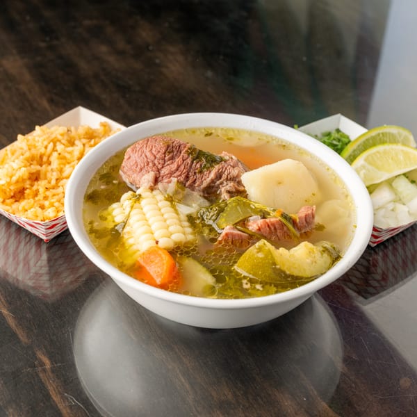 Hearty beef soup with corn, carrots, and potatoes, served with rice and lime wedges.