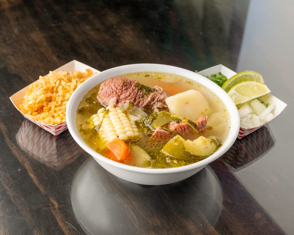 Hearty beef soup with corn, carrots, and potatoes, served with rice and lime wedges.