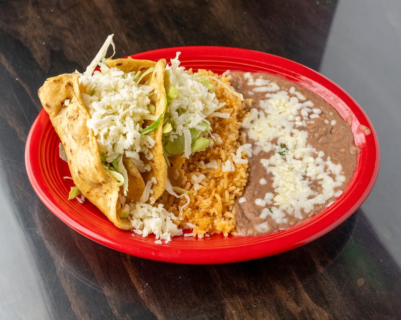 Two crispy tacos with lettuce and cheese, served with rice and refried beans on a red plate.